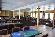 The image shows an empty restaurant or dining area with numerous tables and chairs, decorative tabletops, and large windows providing natural light.