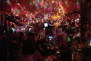 The image shows a bustling bar scene with colorful string lights creating a vibrant, cozy atmosphere.