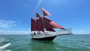 A sailboat with red sails is navigating the sea under a clear blue sky, with several people visible on deck.
