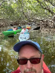 A group of people are kayaking in a mangrove forest with the forefront showing a selfie of a man wearing sunglasses and a baseball cap.
