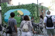 A group of people with bicycles are pausing to look at a vibrant mural of a parrot on an urban building.