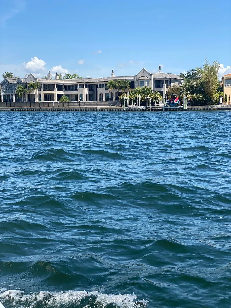 The image shows a view of a large, sprawling waterfront house seen from across a body of choppy blue water under a clear sky.
