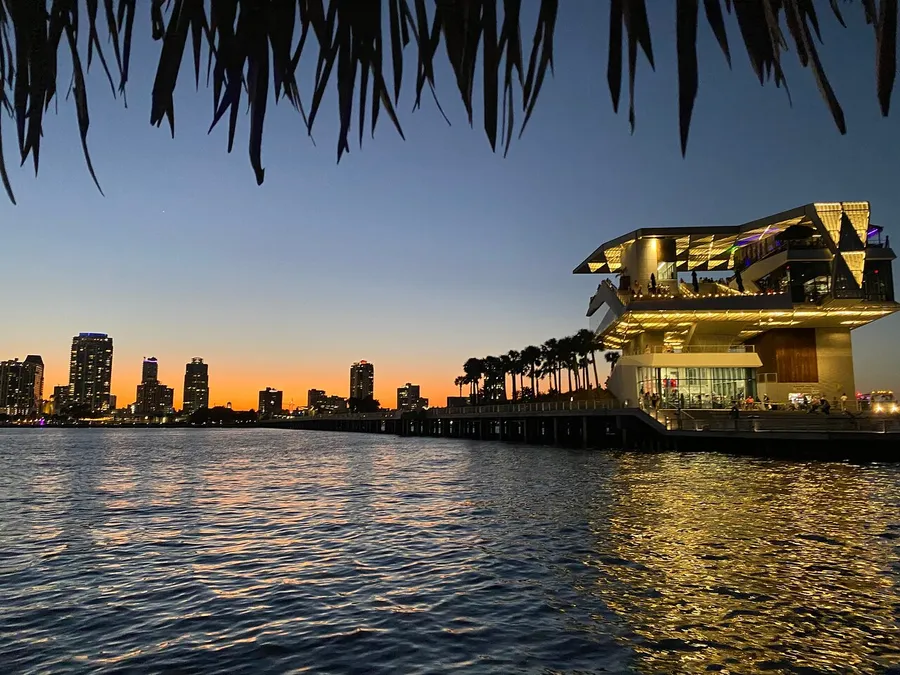 The image captures a modern waterfront building illuminated at twilight with a silhouette of palm trees and a city skyline against an orange-hued sky.