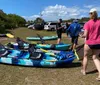 People prepare for a kayaking activity with several colorful kayaks laid out on the grass near parked vehicles under a sunny sky