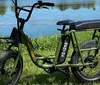 An olive green electric bike with chunky tires is parked on grass against a backdrop of calm water