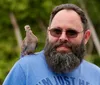 A smiling man with a beard is wearing sunglasses and a blue T-shirt while a dove perches on his shoulder