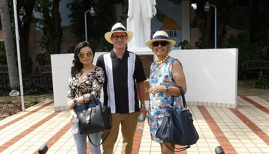Three people wearing sun hats and sunglasses are smiling for the camera while standing outdoors with tropical foliage in the background.