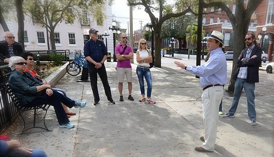 A man in a fedora hat is speaking to a group of attentive people outdoors, possibly conducting a tour or giving a presentation.