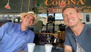 Two smiling men are taking a selfie in a café, with one wearing a straw hat and the other in a casual t-shirt, and a Corona beer sign is visible in the background.