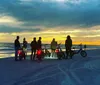 A group of people with bicycles are gathered on a beach at sunset silhouetted against a vibrant sky