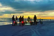 A group of people with bicycles are gathered on a beach at sunset, silhouetted against a vibrant sky.