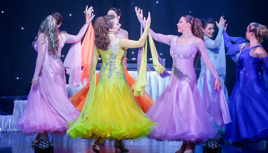 This image shows a group of performers in colorful, sparkling dresses dancing on stage with an air of joy and celebration.
