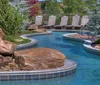 The image shows a serene outdoor lazy river pool flanked by sun loungers and surrounded by lush landscaping