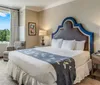 The image depicts a neatly arranged bedroom with a large bed featuring an ornate blue headboard crisp white linens a dark blue decorative throw two bedside tables with lamps and a window offering a view of greenery outside