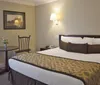 This image shows a neatly arranged hotel room with a patterned bedspread a wooden headboard a small seating area with a table and a framed picture on the wall
