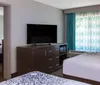 The image shows a neatly arranged modern hotel room with a large bed a work desk and a flat-screen TV