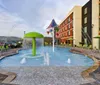 The image showcases an outdoor water play area with a colorful splash pad and seating arrangements situated next to a modern multi-story hotel building