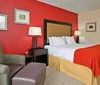 This image shows a neatly arranged hotel room with a king-sized bed a bold red accent wall framed artwork an armchair with an ottoman and a workspace with a desk and chair