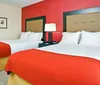 This image shows a neatly arranged hotel room with a king-sized bed a bold red accent wall framed artwork an armchair with an ottoman and a workspace with a desk and chair