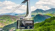 A cable car glides above a lush green forest with picturesque mountains in the background under a partly cloudy sky.