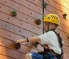 A young person wearing a yellow helmet is focused while climbing a wooden wall with green handholds