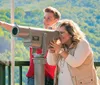 A man and a woman are with a young child who is using a viewing telescope on a balcony overlooking a lush green mountainous landscape with buildings nestled among the trees