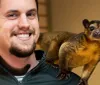 A smiling man poses with a kinkajou perched on his shoulder