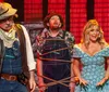 The image shows a lively group of performers on stage likely from a Hatfield  McCoy-themed dinner show with some playing musical instruments and others singing or acting evoking a sense of fun and entertainment