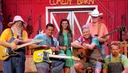 A group of six performers, some holding guitars and one with a microphone, are posing on a stage decorated to resemble a barn with a sign reading 
