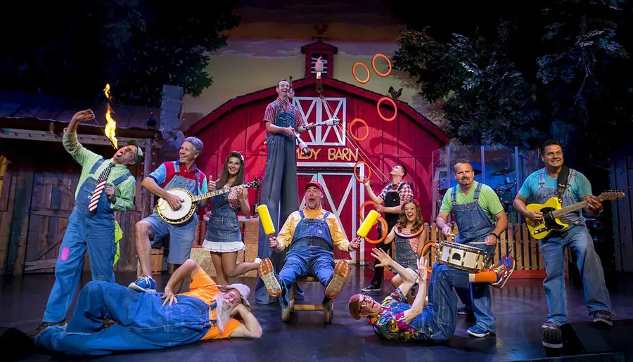 A lively group of performers engages in various entertaining activities such as playing instruments, juggling, and comedy in a barn-themed stage setting.
