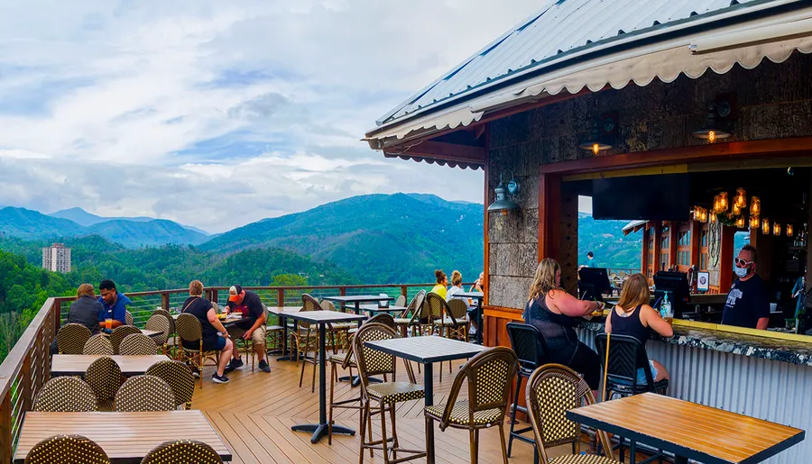 People are enjoying food and drinks at an outdoor mountain-view bar with lush green hills in the background.