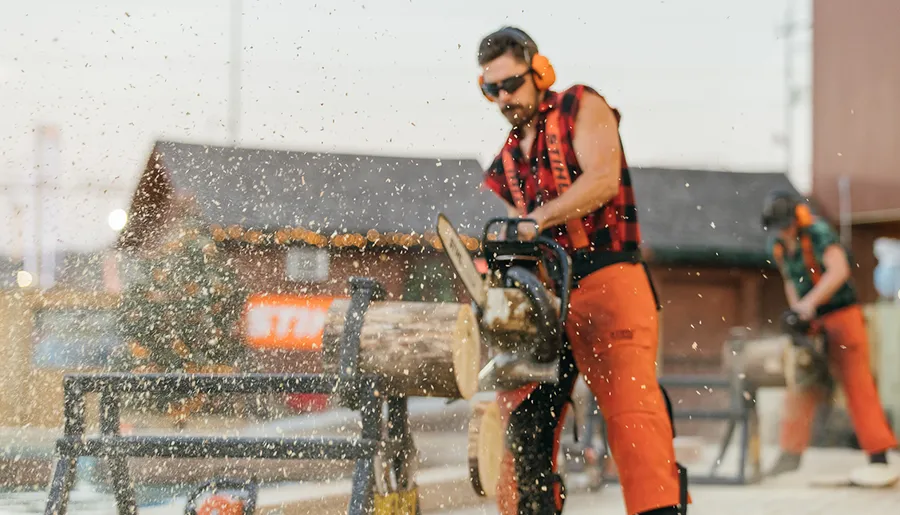 A person wearing safety gear is operating a chainsaw to cut a log, sending wood chips flying into the air.