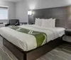 The image shows a modern hotel room with a neatly made king-size bed a patterned carpet a wall-mounted TV a side chair an air conditioner and a simple decor
