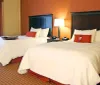 The image shows a hotel room with two neatly made queen-sized beds a nightstand with a lamp and a warm color scheme