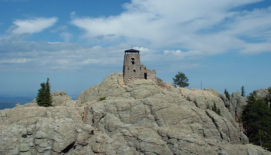 A stone lookout tower sits atop a rugged rocky outcrop against a backdrop of blue skies dotted with clouds.