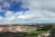 The image shows a panoramic view of a rugged landscape with layered rock formations, green valleys, and a vast sky dotted with clouds.