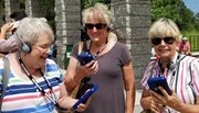 Three smiling women are using audio guide devices likely on an outdoor tour.