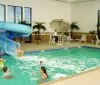 An indoor pool area with a waterslide and people enjoying the water while two individuals sit nearby under an umbrella