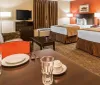 The image shows a neatly arranged double hotel room with two beds a diningwork table set for two and a cozy seating area exuding a warm and inviting ambiance