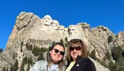 Two people are posing for a photo in front of the Mount Rushmore National Memorial, which features the carved faces of four U.S. presidents.