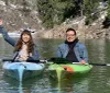 Two people are smiling and enjoying a kayak trip on a tranquil lake with a backdrop of a forested hillside partially covered in snow