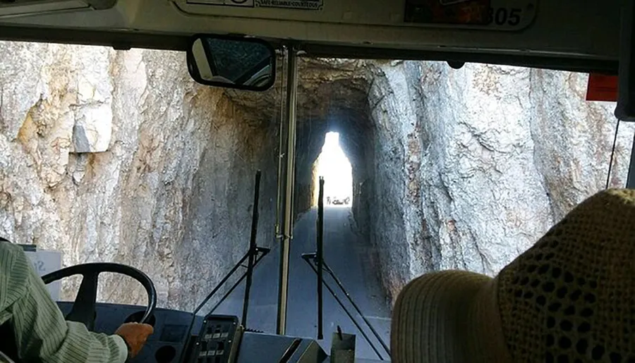 The image shows the interior of a bus as it is about to exit a narrow rock tunnel, with daylight visible at the end of the passage.
