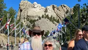 A couple poses for a photo in front of the iconic Mount Rushmore National Memorial, with a walkway lined by various flags.