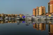 The image depicts a tranquil marina at dusk, with still waters reflecting luxury boats and the surrounding upscale buildings under a fading sky.