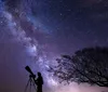 A group of silhouetted people including children and adults are stargazing with telescopes under a starry night sky