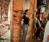 A group of people is attentively listening to a woman speaking in a rustic alleyway with exposed brick and distressed surfaces