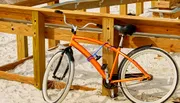An orange bicycle is casually leaned against a wooden railing on a sandy beach under bright sunlight.