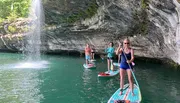 A group of people are paddleboarding near a waterfall and a rocky cliff.