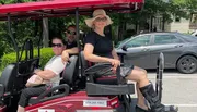 Three people are smiling and seated in a red golf cart parked on a city street on a sunny day.