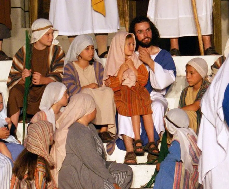A group of people, dressed in biblical-era costumes, appears to be participating in a theatrical or reenactment scene with a man in the center speaking to a child on his lap.